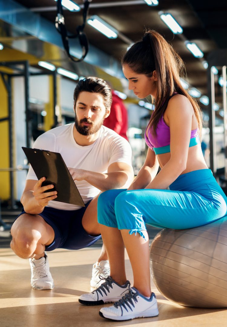 Young healthy active woman sitting on the gym ball and consulting with a  personal trainer about an exercise plan.